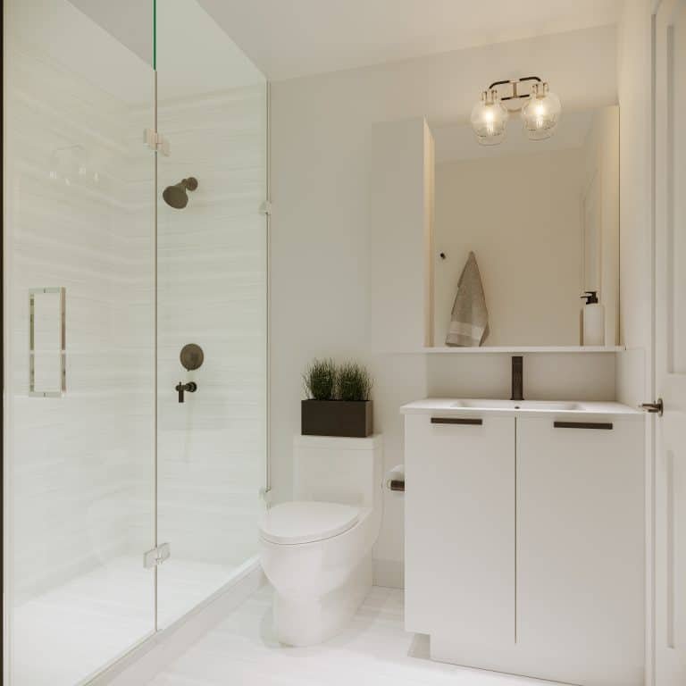 A modern bathroom in new GTA homes showcases a glass-enclosed shower with a black showerhead, a white vanity with black faucet, and a mirror above. A towel hangs neatly to the right, complemented by a potted plant on the shelf behind the toilet. Bright lighting enhances this stylish space.
