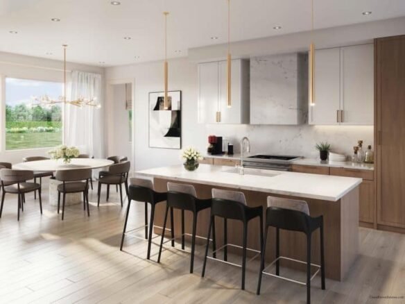 A modern, light-filled kitchen at Clarehaven Estates features a large island with wooden cabinets, white countertops, and four black barstools. Pendant lights hang above the island. A dining table with six chairs sits to the left, near sliding doors that open to a garden view.