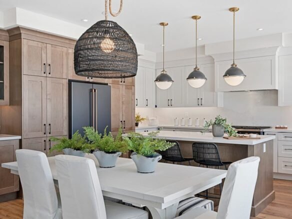 Clarehaven Estates presents a modern kitchen and dining area featuring light wood cabinetry, a white island with black seating, and three pendant lights hanging above. A woven black chandelier is suspended over a white dining table with cushioned chairs and potted plants as a centerpiece.