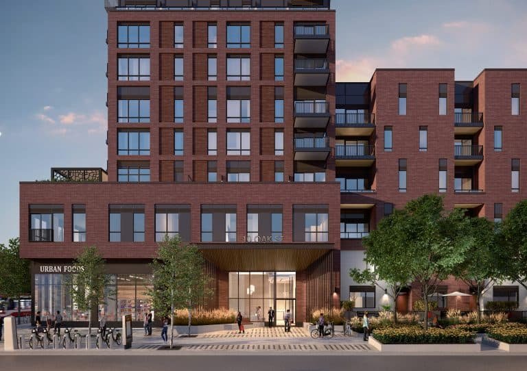 A modern, multi-story red brick building with balconies at various levels showcases the allure of pre-construction homes. Ground-floor shops and large windows invite passersby walking on the tree-lined sidewalk. The sky is clear with a soft pinkish hue, enhancing the charm of new homes in GTA.