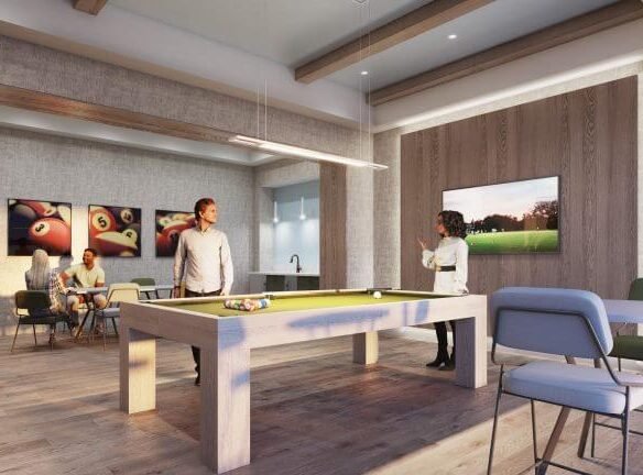 A modern recreational room in a pre-construction home features a pool table at the center. Two people are playing, while two others are seated at a table. The space boasts large windows, bright lighting, and a wall-mounted TV displaying an outdoor scene.