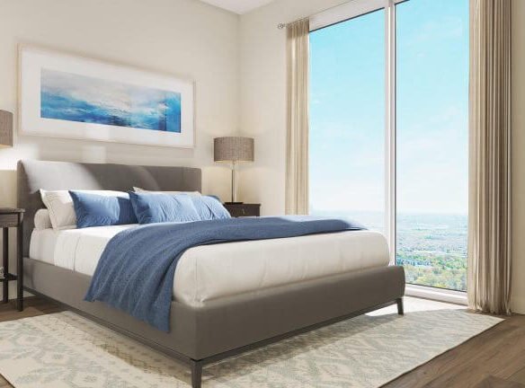 The modern bedroom in this new GTA home features a large bed with a gray headboard and chic white and blue bedding. Two nightstands and lamps flank the bed, while a large window with beige curtains frames views of the landscape. Framed artwork adorns the wall above.