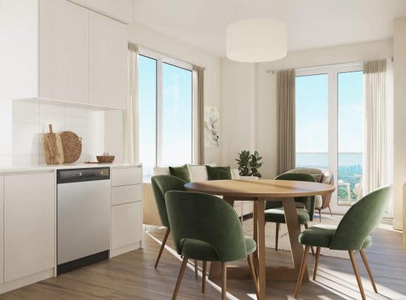 This bright, modern dining area could be yours in one of the new condos by SG Homes. Picture a round wooden table with green velvet chairs basking in sunlight streaming through large windows and a glass door, illuminating white cabinetry and wooden floors while a potted plant sits elegantly in the corner.