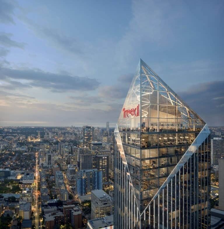 An aerial view of a skyscraper with a distinctive illuminated triangular glass top, prominently displaying the red lettering "free'd". The cityscape below features various buildings and residences, with a street bustling with traffic, extending into the twilight horizon.