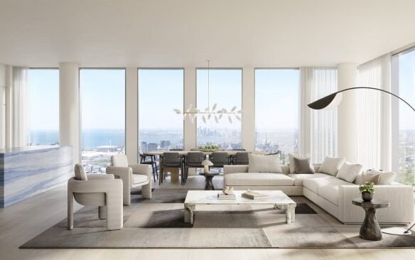 A modern, spacious living room at Freed Residences boasts large floor-to-ceiling windows offering a panoramic city view. The room has a sectional sofa, two armchairs, a marble coffee table, and a unique arc floor lamp. The adjacent kitchen features a blue marbled island.