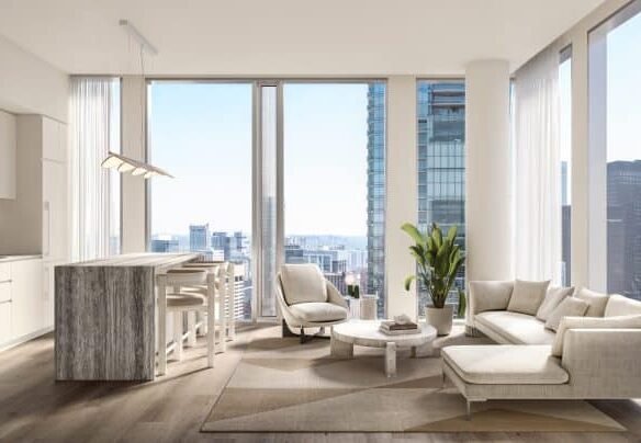 A modern, bright, and spacious living room at Freed Residences with floor-to-ceiling windows showcasing a city skyline. The room features a beige sectional sofa, a round coffee table, a small dining table with four chairs, neutral-toned decor, and light hardwood floors.