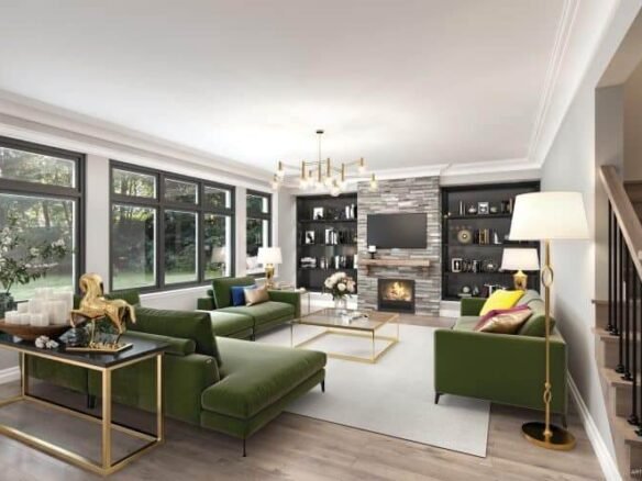 A modern living room in a pre-construction home features a large green sectional sofa, gold-accented furniture, and a stone fireplace. Built-in shelves with decor items surround the fireplace. Expansive windows let in natural light, while a chandelier and floor lamp illuminate the space.