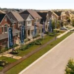 Aerial view of a residential neighborhood featuring pre-construction homes with modern two-story designs. Each house boasts a front yard and driveway. Tall banners line the tree-lined street, running parallel to these new homes in GTA, all under a clear sky.
