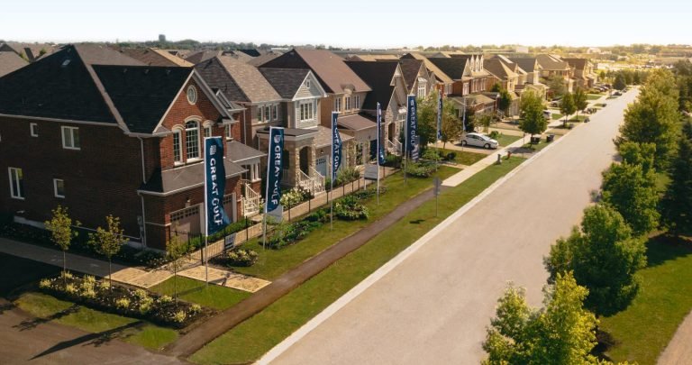 Aerial view of a residential neighborhood featuring pre-construction homes with modern two-story designs. Each house boasts a front yard and driveway. Tall banners line the tree-lined street, running parallel to these new homes in GTA, all under a clear sky.
