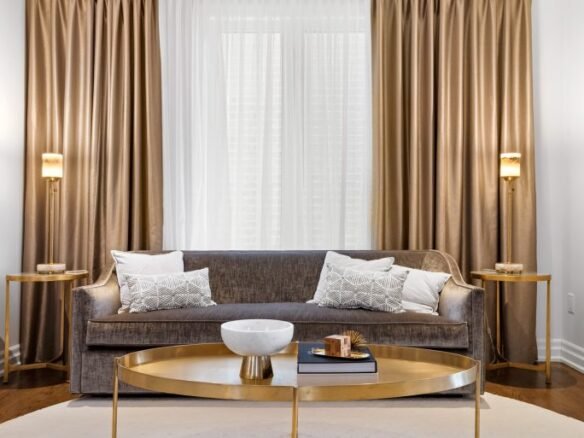 A modern living room by Sg Homes features a gray couch adorned with white and patterned pillows. Two gold side tables with lamps complement a large round coffee table. Beige curtains frame sheer white drapes, enhancing the elegant feel of these pre-construction homes.