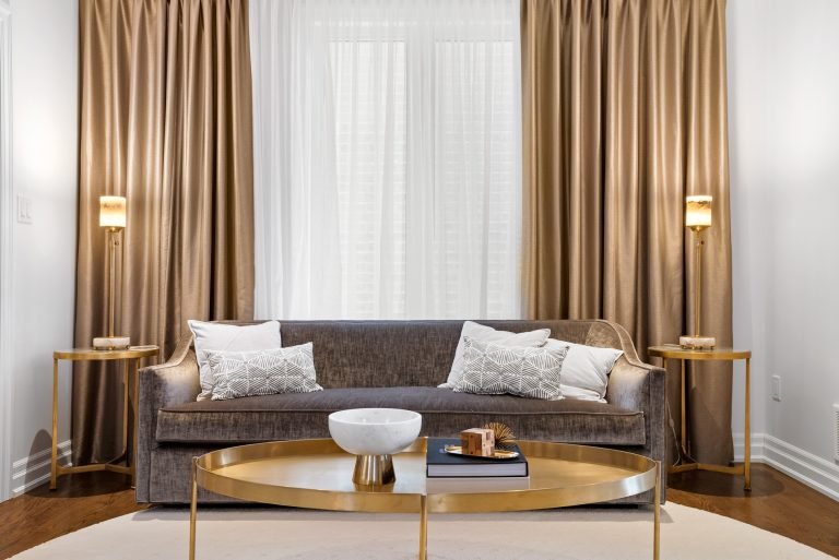 A modern living room by Sg Homes features a gray couch adorned with white and patterned pillows. Two gold side tables with lamps complement a large round coffee table. Beige curtains frame sheer white drapes, enhancing the elegant feel of these pre-construction homes.