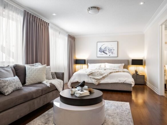 A modern bedroom in one of the new condos features a large bed with white bedding and a faux fur throw. A beige sofa with cushions sits in the foreground, while a round coffee table holds decorative items. Two side tables with black lamps flank the bed, and natural light floods the inviting space.