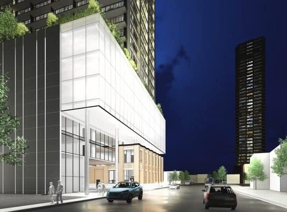 A nighttime view of a modern city street showcases tall buildings in the foreground. A glass-paneled structure with rooftop plants hints at new condos, as two cars glide along the street. Pedestrians stroll nearby, while another towering building stands proudly in the background.