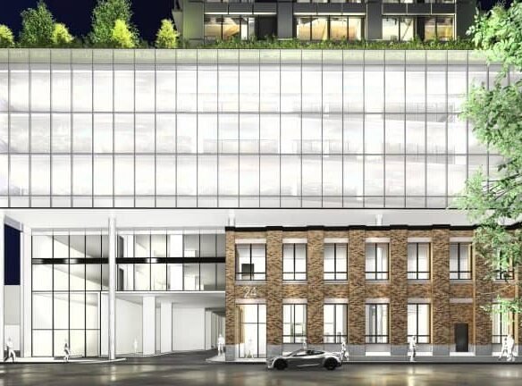 Modern building at night blending glass and brick architecture. The lower section features brick with tall windows, while the sleek, glass-fronted upper part exudes elegance. A few trees adorn the rooftop of these new homes in GTA. A car is parked in front as people stroll by.