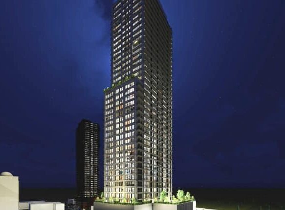 Night view of a tall, illuminated skyscraper with numerous lit windows surrounded by smaller buildings. The clear, dark sky enhances the building's lights, while new condos rise alongside. Trees frame the base of this modern marvel, offering a glimpse into urban living at its finest.