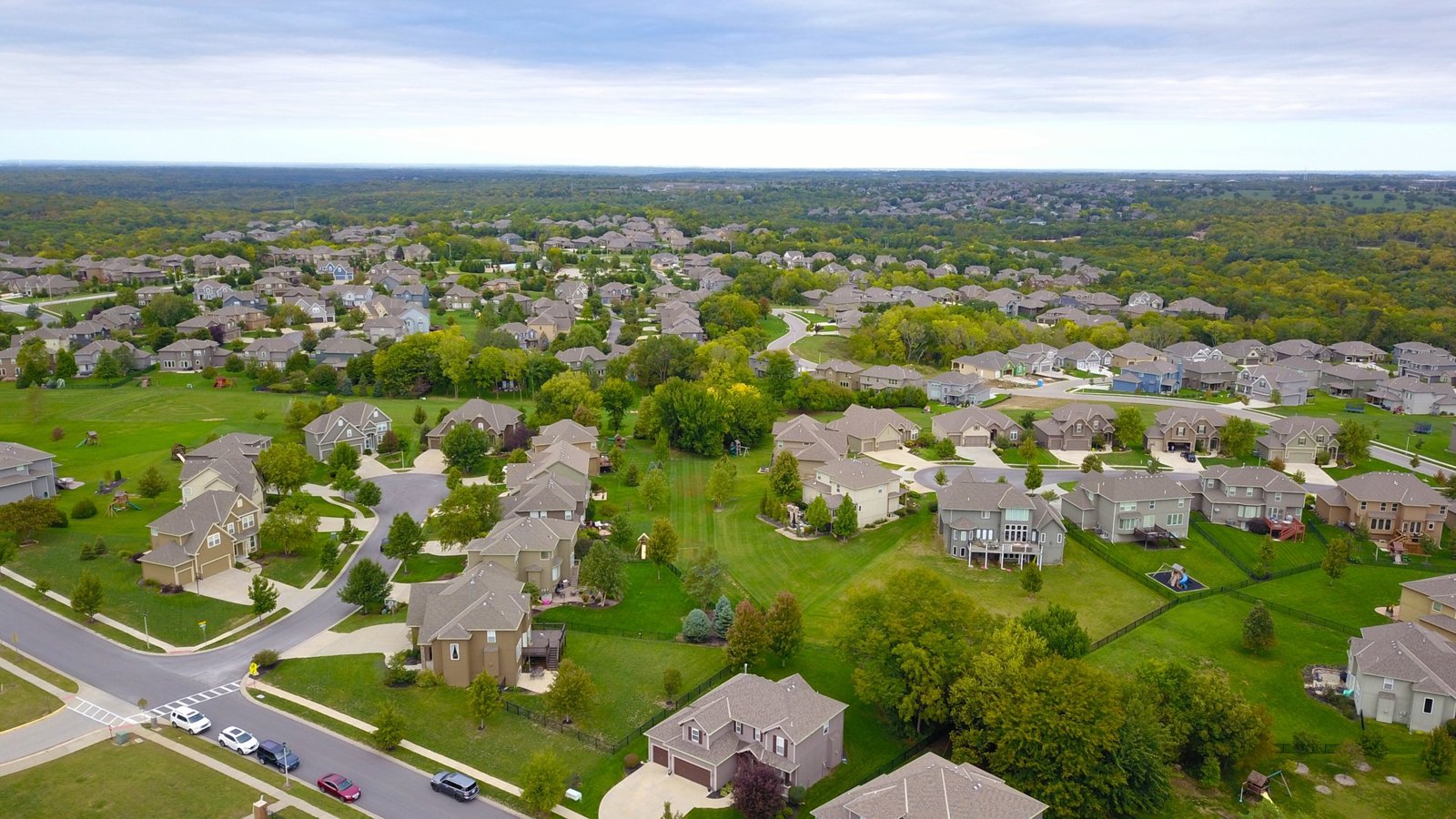 Aerial view of a suburban neighborhood with rows of houses and new homes surrounded by green lawns and trees. A network of roads connects the residences, extending to a forested area in the background under a cloudy sky, showcasing the blend of nature and development.