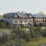 A row of modern townhouses with sloped roofs is situated behind a street lined with trees and grass. These new homes offer a fresh take on urban living. In the foreground, there is a small parking area and a few traffic cones. The sky is clear.