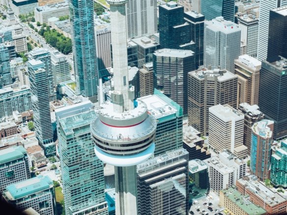 Aerial view of downtown Toronto featuring the CN Tower in the center. The bustling landscape is filled with modern skyscrapers and newly constructed condos, showcasing a vibrant urban vibe. People are visible on the CN Tower's observation deck, enjoying this expansive cityscape.