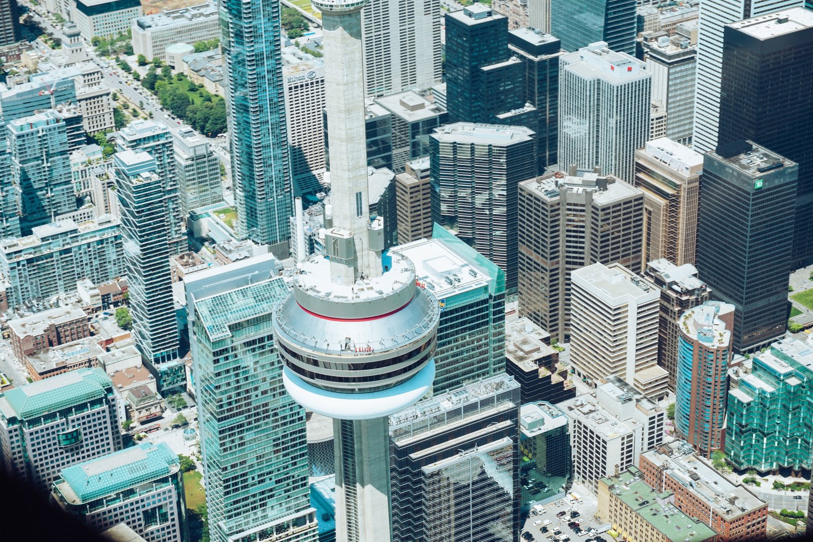 Aerial view of downtown Toronto featuring the CN Tower in the center. The bustling landscape is filled with modern skyscrapers and newly constructed condos, showcasing a vibrant urban vibe. People are visible on the CN Tower's observation deck, enjoying this expansive cityscape.