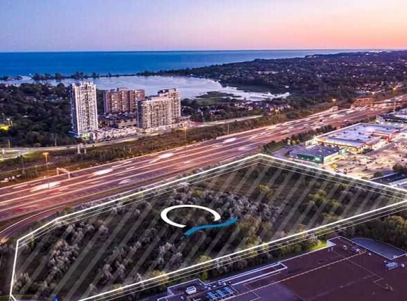 Aerial view of a cityscape at dusk, featuring a highway with mesmerizing light trails, surrounding buildings, and pre-construction homes outlined by white lines. In the background, there's a large body of water and new condos in the GTA among residential high-rises.