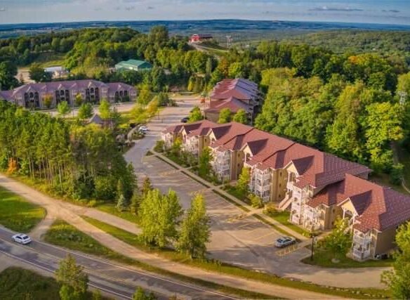 Aerial view of a residential area featuring rows of large, multi-story buildings with reddish-brown roofs, surrounded by lush green trees, near the Carriage Country Club, under a clear sky.