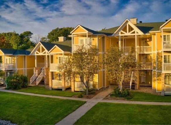 Aerial view of the Carriage Country Club apartment complex featuring multiple three-story buildings with balconies and external staircases, surrounded by lush greenery and a well-maintained lawn, bathed in warm sunlight.