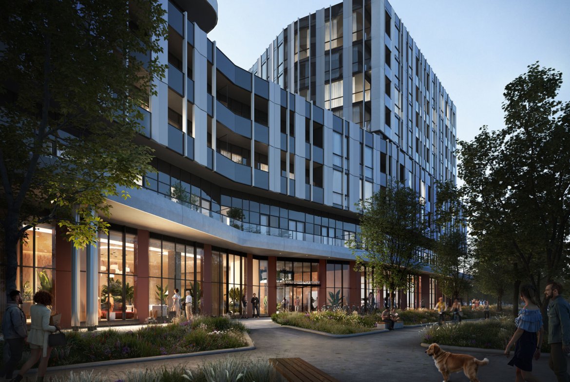 The Clove is a modern multi-story building with a glass and metal exterior, featuring ground-level retail spaces lit warmly inside. People are walking on the pathway outside, with trees and plants lining the route. A person is walking a dog in the foreground.