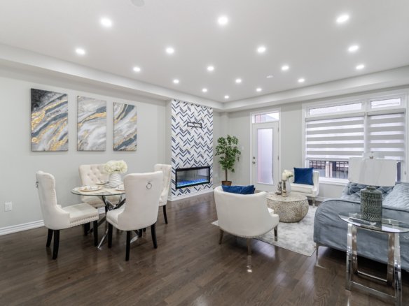 A modern living and dining area featuring white upholstered chairs, glass-top dining table, wall art, and a fireplace with chevron-patterned tiles. Large windows and recessed lighting enhance the spacious, elegant room, perfect for embracing an empty nest new lifestyle with its gray sofa and coffee table.