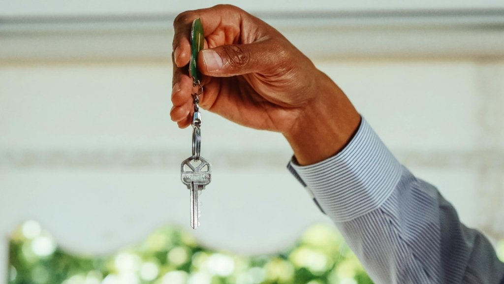 A person wearing a light blue, long-sleeve shirt is holding a keychain with two keys attached, perhaps symbolic of navigating the real estate market. The hand is positioned at the top center of the image, and the background is blurred with soft, natural light.