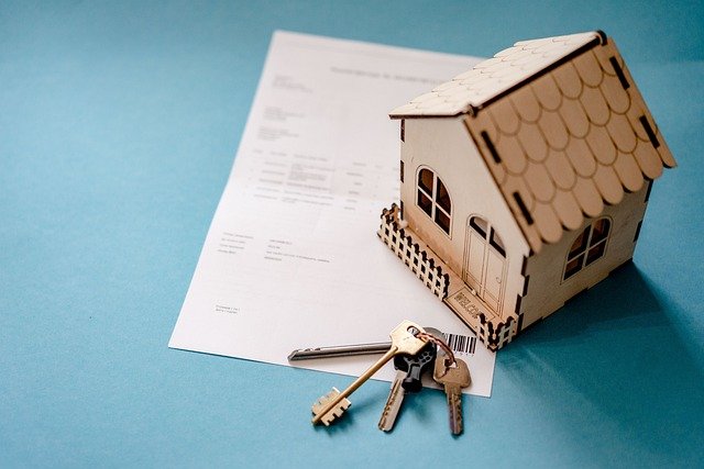 A small wooden model of a house sits on top of a document, likely related to the complexities of real estate or financial contracts, with a set of three metal keys attached to a keyring placed in front. The background is a plain blue surface. This scene captures the essence of home construction in Canada.