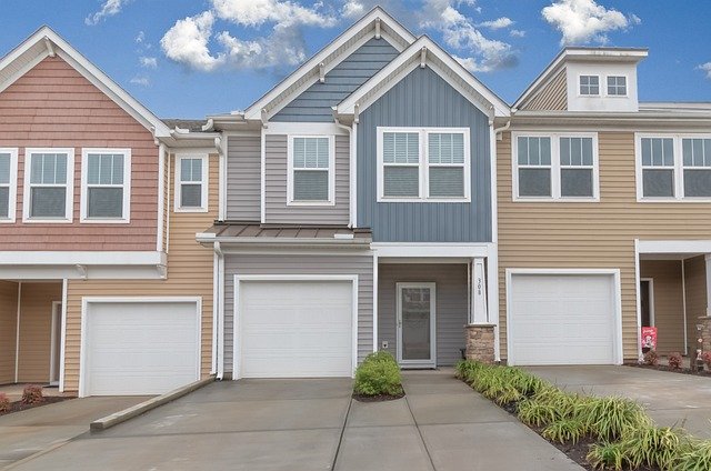 A row of modern townhouses with three visible units, each equipped with a garage and a front entrance. The exteriors feature different shades of brown, gray, and blue siding. Embracing the Empty Nest: Transformations for a New Lifestyle can be seen in the carefully manicured shrubs and plants lining the front yards under a partly cloudy sky.