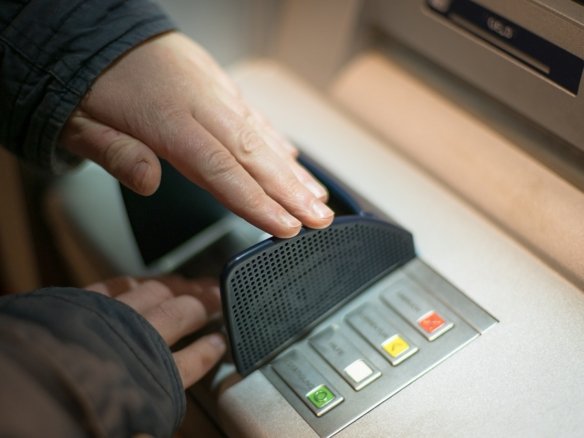 A person discreetly shields the keypad with their hand while entering a PIN at an ATM, perhaps contemplating recent RRSP changes for future homeownership. The machine's colorful buttons glow under the dim lighting of this indoor setting, its screen hovering above like a silent guardian.