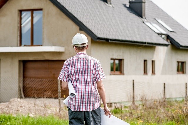 A person in a white hard hat and checkered shirt, possibly consulting a homeowner's guide, holds architectural plans while facing a two-story house with a sloped roof and several windows. The house is partially obscured by a fence and grass, hinting at ongoing renovations.