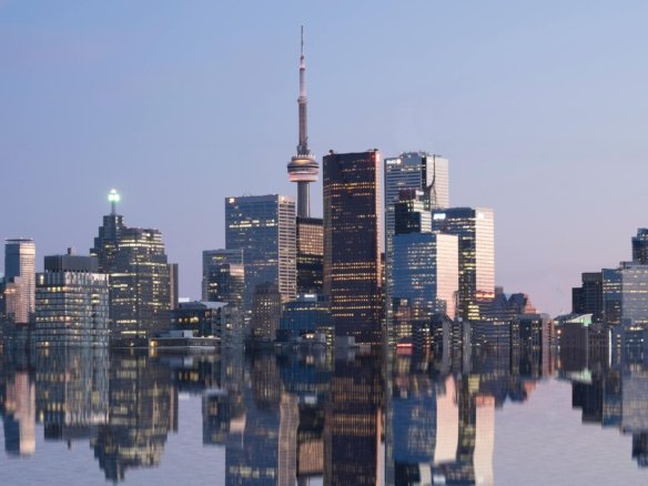 Skyline of a city with modern skyscrapers reflecting on a calm body of water during twilight. A prominent tower stands at the center. The buildings' lights and the faint pink sky create a serene urban scene.