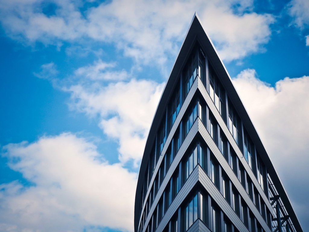 A modern building with a sharp, triangular roof and glass windows reaches upwards against a backdrop of a partly cloudy blue sky. The structure's sleek lines and reflective surfaces create a striking, contemporary architectural scene.