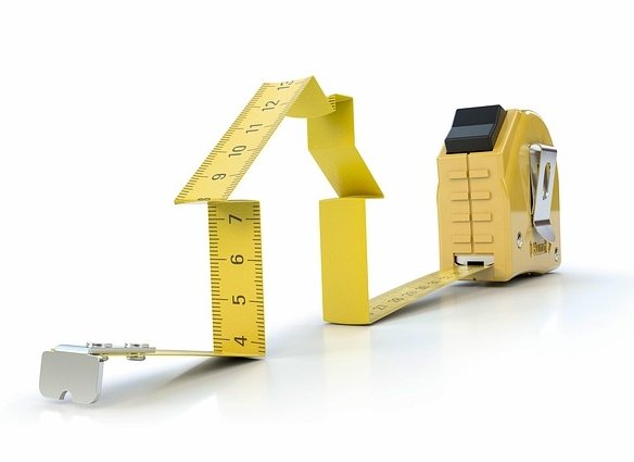 A yellow tape measure is creatively bent to form the shape of a house, symbolizing endless renovation tips. The tape extends from its metal case, resting on a white surface, ready to inspire your next home project.