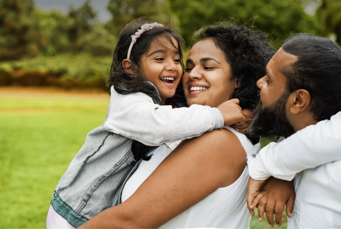 A joyful family is outdoors at Caledon Club, with a young girl hugging her smiling mother. The father and another child are beside them, all enjoying time together in a green park.