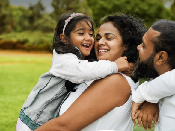 A joyful family is outdoors at Caledon Club, with a young girl hugging her smiling mother. The father and another child are beside them, all enjoying time together in a green park.