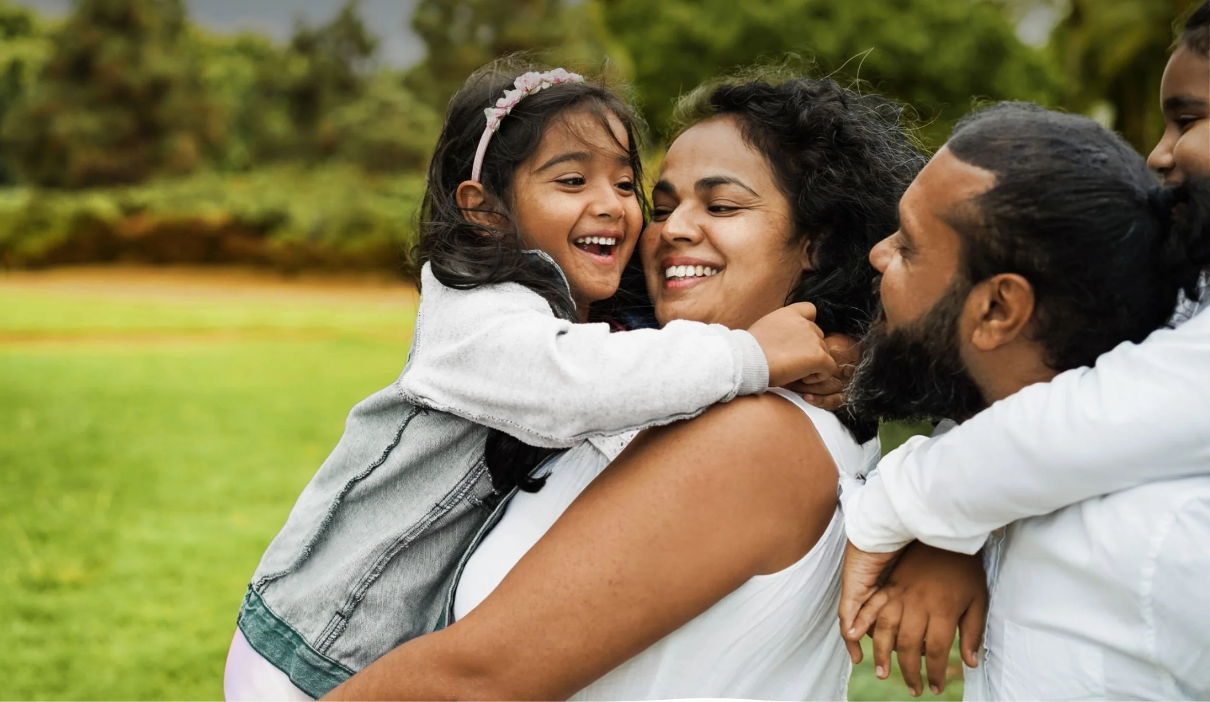 A joyful family is outdoors at Caledon Club, with a young girl hugging her smiling mother. The father and another child are beside them, all enjoying time together in a green park.