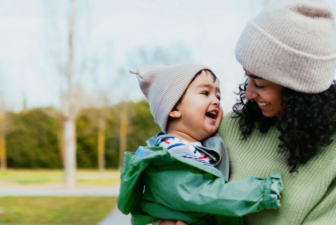 A woman in a green sweater and beige beanie holds a smiling young child in a green jacket and matching beige hat. Outdoors on a clear day, they happily look at each other, enjoying a perfect Caledon Club afternoon together.