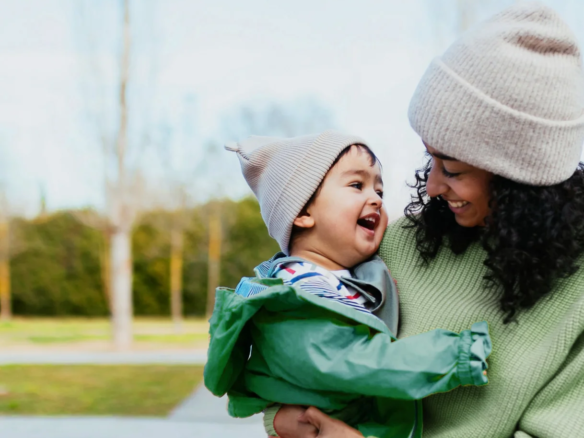 A woman in a green sweater and beige beanie holds a smiling young child in a green jacket and matching beige hat. Outdoors on a clear day, they happily look at each other, enjoying a perfect Caledon Club afternoon together.